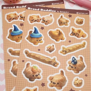 May include: A collection of stickers featuring adorable bread-themed animal designs, including cats and dogs. The stickers are on a grid-patterned background, with some wearing hats. A roll of tape and pens are also visible.