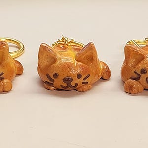 Könnte beinhalten: Drei orangefarbene Katzen-Schlüsselanhänger mit goldfarbenen Ringen. Jeder Schlüsselanhänger hat ein lächelndes Gesicht mit dunkelbraunen Augen und Schnurrhaaren. Die Schlüsselanhänger liegen auf einer hellen Oberfläche.