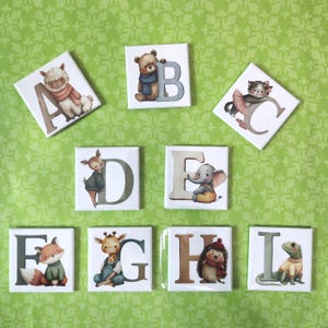 May include: A set of nine square tiles featuring illustrations of animals paired with letters A through I. Each tile displays a letter in a soft color alongside a character: a llama, bear, cat, deer, elephant, fox, giraffe, hedgehog, and lizard.