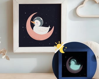 Puede incluir: Una impresión enmarcada con una ilustración de un pato con un casco espacial, sentado en una luna creciente rosa sobre un fondo azul oscuro estrellado. El marco es blanco. Se muestra una versión más pequeña y brillante de la impresión debajo.