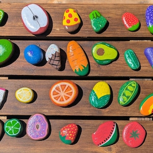 May include: A collection of hand-painted rocks depicting various fruits and vegetables. The assortment includes a lime, apple, kiwi, blueberry, carrot, avocado, and more. Each rock is painted with vibrant colors and detailed designs, creating a playful and artistic display.