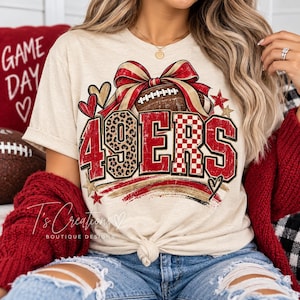 Könnte beinhalten: Ein cremefarbenes T-Shirt mit einem Grafikdesign, das den Text 49ERS in Rot, Leopardenmuster und Karomuster zeigt. Das Design umfasst einen Football, eine große rot-goldene Schleife, Herzen und Sterne. Das T-Shirt hat einen geknoteten Saum.