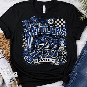 May include: A black short-sleeve t-shirt featuring a graphic print of a blue and white coiled rattlesnake. The design includes the text "RATTLERS" and "PRIDE" in blue and white, accented with blue bows, checkered patterns, and small heart shapes.