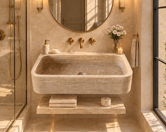 May include: A wall-mounted beige stone sink with a rectangular basin, featuring brass fixtures, a round mirror with a gold frame, two vertical wall sconces, a stone shelf with folded towels, and a small vase of white flowers on a marble surface.