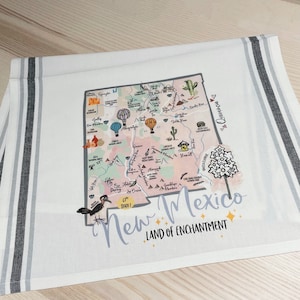 May include: A white tea towel with a black and white striped border features a colorful illustrated map of New Mexico. The map includes various landmarks, cities, and the text "New Mexico, Land of Enchantment".
