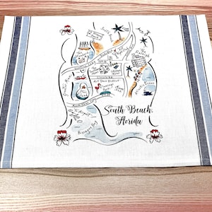 May include: A white tea towel with blue stripes on the sides. The center features a hand-drawn map of South Beach, Florida, with various landmarks and points of interest labeled. The map is surrounded by a blue wave design and red hibiscus flowers.