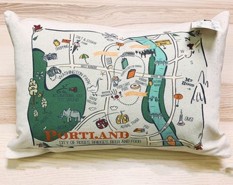 May include: A white cushion featuring a green and blue map of Portland, Oregon. The map highlights landmarks such as the International Rose Test Gardens, Goose Hollow, and the Aerial Tram. The text "Portland, City of Roses, Bridges, Beer and Food" is printed below the map.