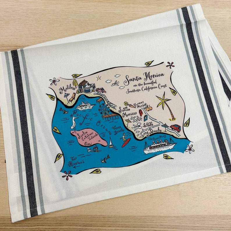 May include: A white tea towel with a blue and pink map of Santa Monica, California. The map shows the Pacific Ocean, Santa Monica Pier, and other landmarks. The text "Santa Monica on the beautiful Southern California Coast" is printed on the towel.