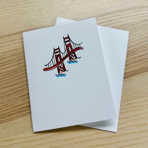 May include: A white card with a red and black line drawing of the Golden Gate Bridge. The bridge is drawn with red lines and black outlines. The water is blue.