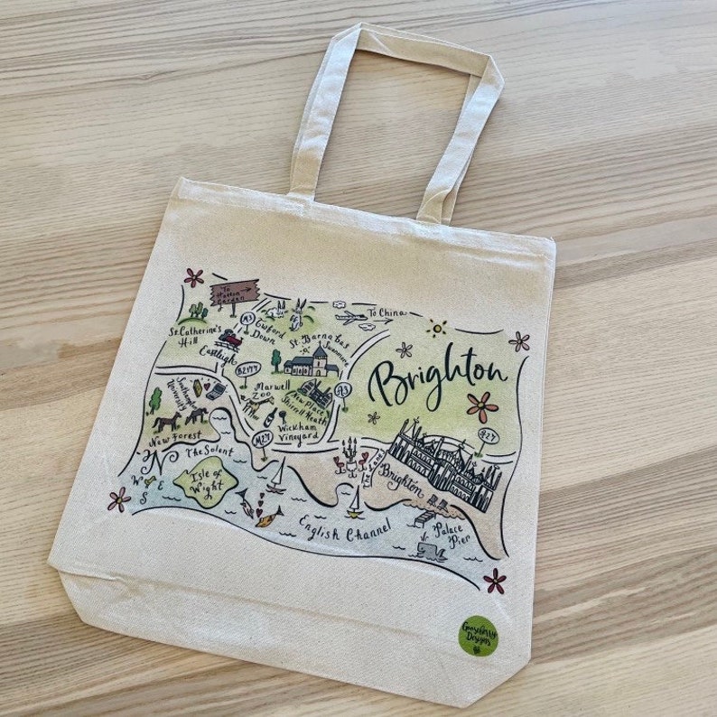 Puede incluir: Una bolsa de tela beige con un mapa dibujado a mano de Brighton, Inglaterra. El mapa presenta puntos de referencia como el Palace Pier, Brighton Beach y el Canal de la Mancha. La bolsa tiene un logotipo verde que dice "Friendly Shops".