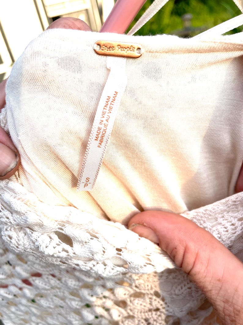 Puede incluir: Primer plano de un vestido color crema con ribete de encaje. El vestido tiene una etiqueta que dice "Free People" y "Hecho en Vietnam". La talla es peque&ntilde;a.