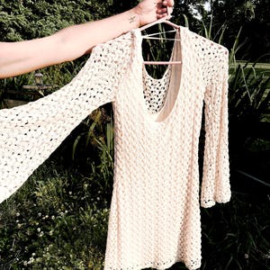 Puede incluir: Un vestido de crochet color crema, de manga larga, con cuello redondo. El vestido est&aacute; colgado de una percha rosa. El patr&oacute;n de crochet es visible en todo el vestido, y las mangas tienen un dise&ntilde;o acampanado.