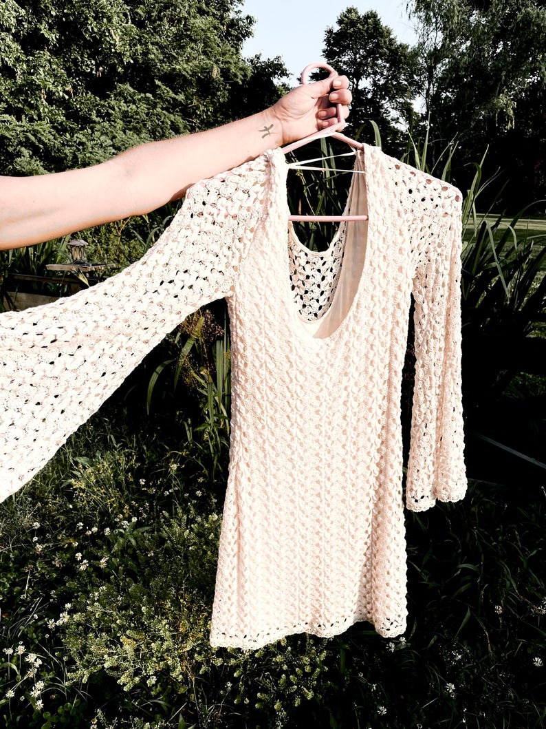 Puede incluir: Un vestido de crochet color crema, de manga larga, con cuello redondo. El vestido est&aacute; colgado de una percha rosa. El patr&oacute;n de crochet es visible en todo el vestido, y las mangas tienen un dise&ntilde;o acampanado.