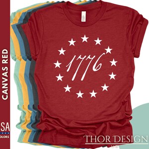Pode incluir: Camiseta de lona vermelha com um c&iacute;rculo branco de estrelas e o texto "1776" no centro. A camiseta &eacute; feita nos Estados Unidos e est&aacute; dispon&iacute;vel em 55 cores.