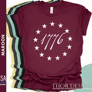 Pode incluir: Uma camiseta de cor vinho com um c&iacute;rculo branco de estrelas que envolve o ano 1776. O texto "THOR DESIGN" est&aacute; impresso na parte inferior da camiseta.