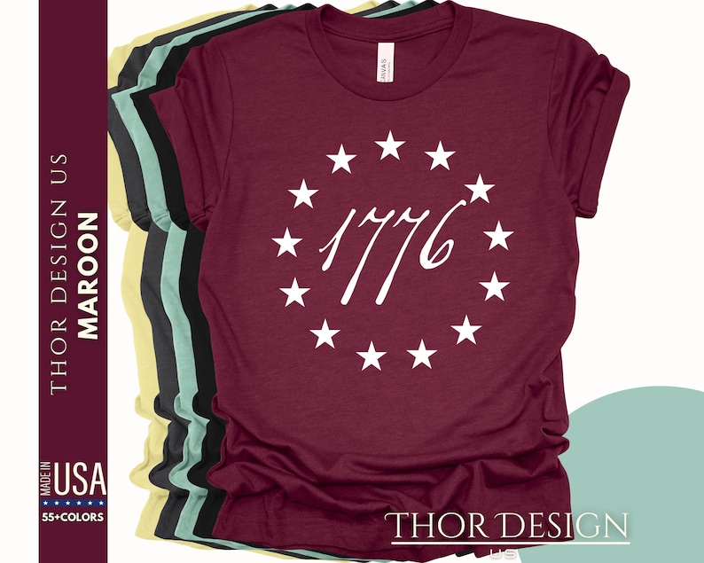 Pode incluir: Uma camiseta de cor vinho com um c&iacute;rculo branco de estrelas que envolve o ano 1776. O texto "THOR DESIGN" est&aacute; impresso na parte inferior da camiseta.