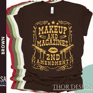 May include: Brown t-shirt with a gold graphic design that says "Makeup and Magazines 2nd Amendment". The graphic features two pistols and a lipstick kiss.