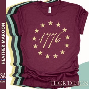 May include: A maroon t-shirt featuring a circle of stars and the number 1776 in gold lettering. The shirt is made from a soft, comfortable material and is available in a range of colours.