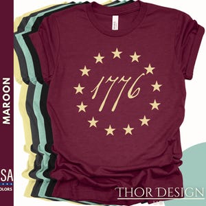 May include: Maroon t-shirt featuring a circle of gold stars and the text "1776" in a gold script font.