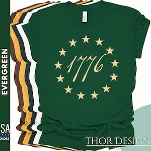 May include: A green t-shirt featuring a gold circle of stars and the text "1776" in a gold script font. The shirt is available in a range of colours.