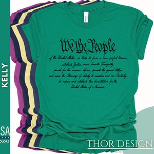 May include: A green t-shirt with the text "We the People" in black cursive font. The text is from the preamble of the United States Constitution. The shirt is folded and stacked with other t-shirts in different colors.