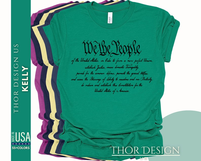May include: A green t-shirt with the text "We the People" in black cursive font. The text is from the preamble of the United States Constitution. The shirt is folded and stacked with other t-shirts in different colors.