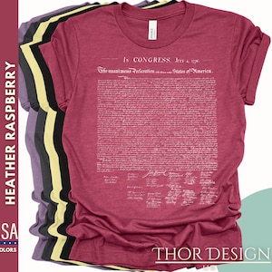 May include: A heather raspberry t-shirt with the full text of the Declaration of Independence printed in white on the front. The text includes the date "July 4, 1776" and the signatures of the signatories. The shirt is shown folded with other t-shirts in different colors.