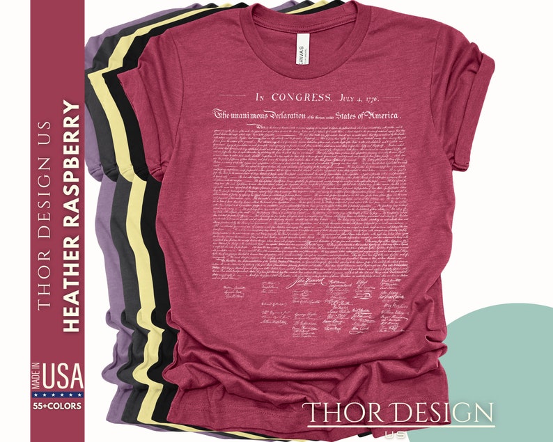 May include: A heather raspberry t-shirt with the full text of the Declaration of Independence printed in white on the front. The text includes the date "July 4, 1776" and the signatures of the signatories. The shirt is shown folded with other t-shirts in different colors.
