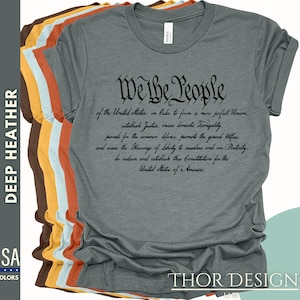 May include: A dark heather grey t-shirt with the text "We the People" and the preamble to the United States Constitution printed in black.