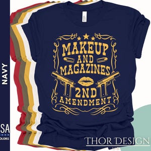 May include: Navy blue t-shirt with a gold graphic design featuring two guns crossed with a lipstick kiss and the text "Makeup and Magazines 2nd Amendment".
