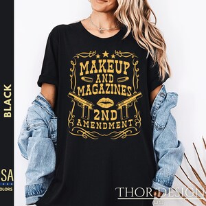 May include: Black t-shirt with a gold graphic design that says "Makeup and Magazines 2nd Amendment". The graphic features two pistols and a lipstick kiss.