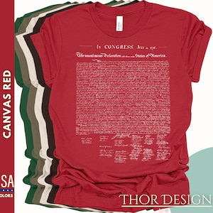 May include: Red t-shirt with a white print of the Declaration of Independence. The text includes the date "July 4, 1776" and the phrase "In Congress, July 4, 1776". The t-shirt is folded and displayed with other t-shirts in different colors.