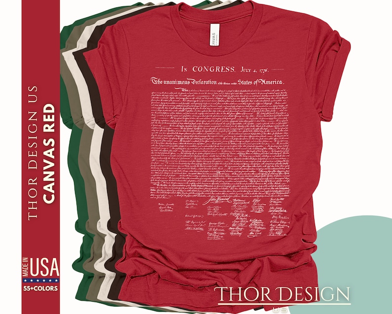 May include: Red t-shirt with a white print of the Declaration of Independence. The text includes the date "July 4, 1776" and the phrase "In Congress, July 4, 1776". The t-shirt is folded and displayed with other t-shirts in different colors.