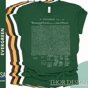 May include: A green t-shirt with the full text of the Declaration of Independence printed on the front in white. The text includes the signatures of the founding fathers. The t-shirt is folded and stacked with other t-shirts in different colors.