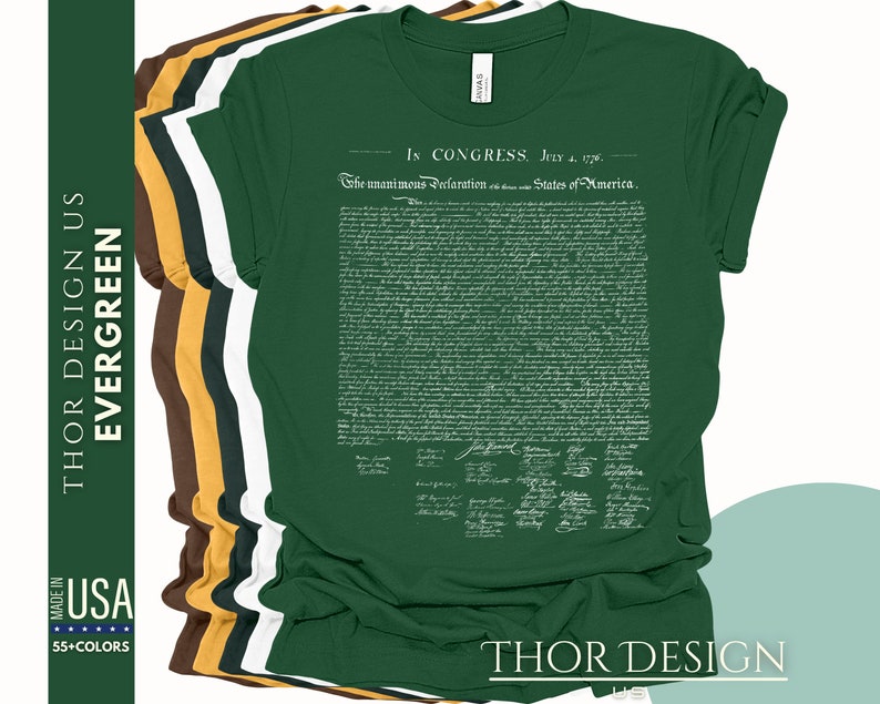 May include: A green t-shirt with the full text of the Declaration of Independence printed on the front in white. The text includes the signatures of the founding fathers. The t-shirt is folded and stacked with other t-shirts in different colors.