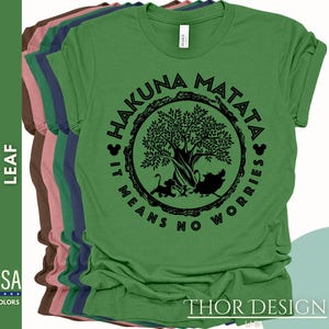 Puede incluir: Camiseta verde con un gr&aacute;fico circular negro que presenta un &aacute;rbol, animales y el texto "Hakuna Matata It Means No Worries".