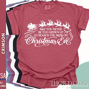 May include: A crimson red t-shirt with a white graphic of Santa Claus flying in a sleigh with reindeer. The text reads "May you never be too grown up to search the skies on Christmas Eve".