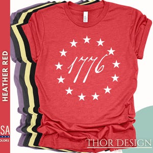 Pode incluir: Camiseta vermelha de mistura com um c&iacute;rculo branco de estrelas e o n&uacute;mero 1776 no centro. O texto "THOR DESIGN" est&aacute; na parte inferior da camiseta.