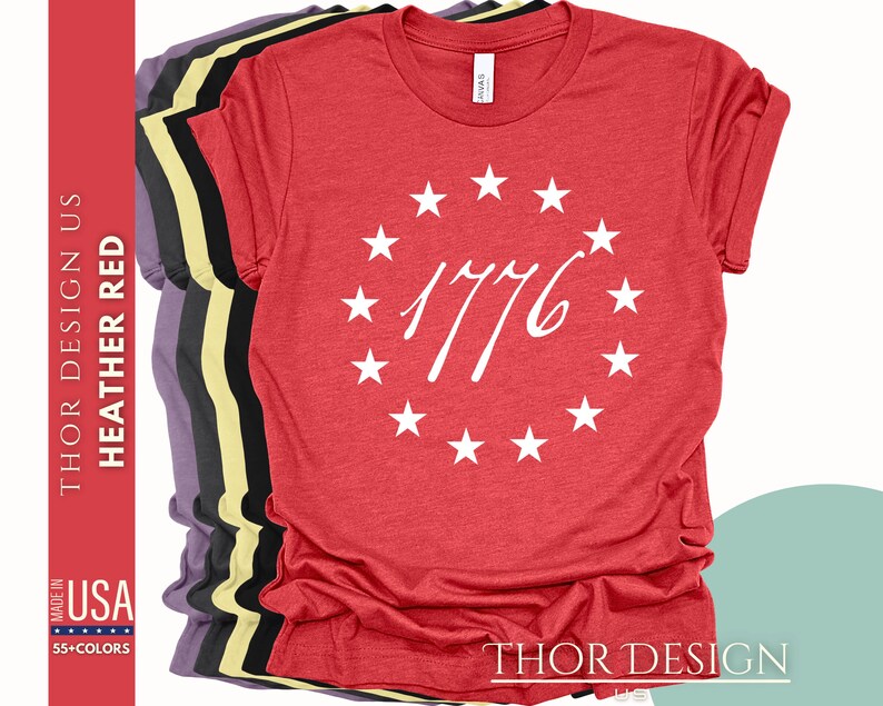 Pode incluir: Camiseta vermelha de mistura com um c&iacute;rculo branco de estrelas e o n&uacute;mero 1776 no centro. O texto "THOR DESIGN" est&aacute; na parte inferior da camiseta.