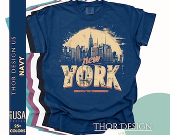 May include: A navy blue t-shirt with a graphic design of the New York City skyline in a vintage style. The text "New York" is in a distressed font and the skyline is in a muted color palette. The t-shirt is made in the USA and is available in 25+ colors.