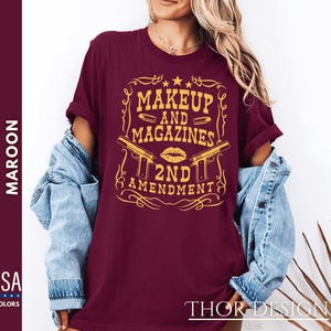 May include: A maroon t-shirt with a gold graphic design that says "Makeup and Magazines 2nd Amendment". The graphic features a stylized gun, lipstick, and bullets.