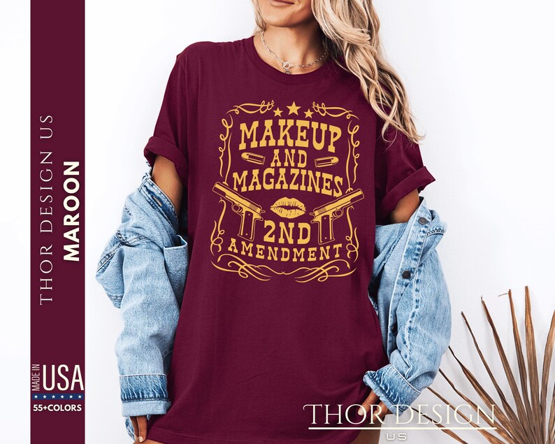 May include: A maroon t-shirt with a gold graphic design that says "Makeup and Magazines 2nd Amendment". The graphic features a stylized gun, lipstick, and bullets.