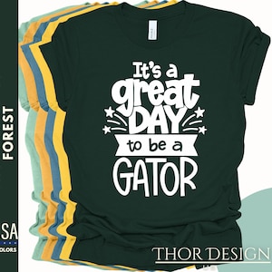 May include: Dark green t-shirt featuring the white text "It's a great DAY to be a GATOR". The image shows a selection of t-shirts in different colours. The brand name "THOR DESIGN US" is visible.