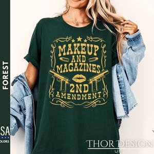 May include: A dark green t-shirt with a gold graphic design featuring two pistols, a lipstick kiss, and the text "Makeup and Magazines 2nd Amendment".