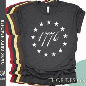 Pode incluir: Camiseta cinza escuro heather com um c&iacute;rculo branco de estrelas que envolve o ano 1776. O texto "THOR DESIGN US" est&aacute; impresso no lado esquerdo da camiseta e "THOR DESIGN" est&aacute; impresso no lado inferior direito da camiseta.