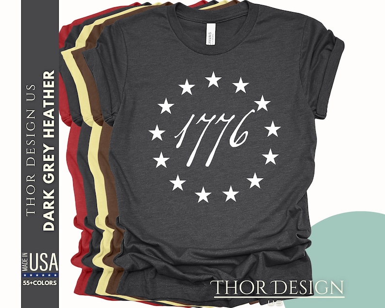 Pode incluir: Camiseta cinza escuro heather com um c&iacute;rculo branco de estrelas que envolve o ano 1776. O texto "THOR DESIGN US" est&aacute; impresso no lado esquerdo da camiseta e "THOR DESIGN" est&aacute; impresso no lado inferior direito da camiseta.