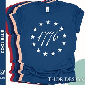 Pode incluir: Uma camiseta azul marinho com um c&iacute;rculo branco de estrelas ao redor do n&uacute;mero 1776. O texto "THOR DESIGN US" est&aacute; impresso na parte inferior da camiseta.