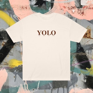 Puede incluir: Camiseta color crema con la palabra "YOLO" en marrón, sobre las palabras "You Only Live Once". La camiseta está hecha de un material suave y se muestra sobre un fondo de graffiti colorido.
