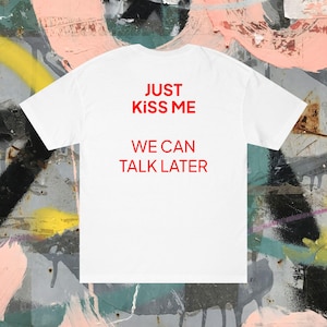 Puede incluir: Camiseta blanca con el texto rojo "JUST KISS ME" encima de "WE CAN TALK LATER". La camiseta se muestra sobre un fondo de graffiti colorido.
