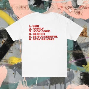 Puede incluir: Camiseta blanca con texto rojo que enumera seis objetivos de vida: Dios, Familia, Verse bien, Ser rico, Tener éxito y Mantenerse privado. La camiseta se muestra sobre un fondo de graffiti colorido.